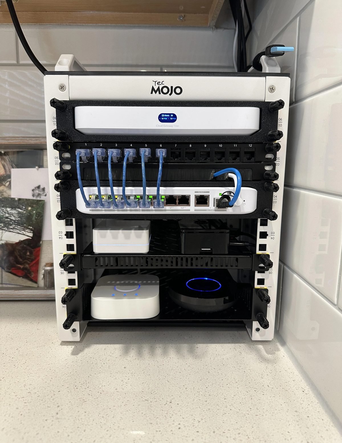 Compact wall-mounted network rack containing a UniFi Cloud Gateway Fiber, a 12-port patch panel and a PoE switch, with blue patch cables neatly dressed through a brush panel